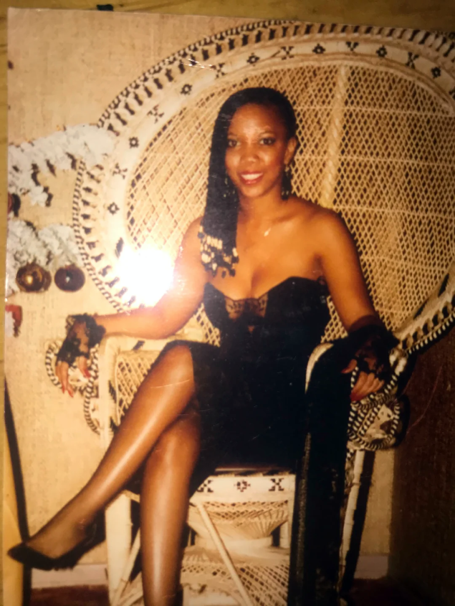 A woman in a black strapless dress sits on a white wicker peacock chair, smiling at the camera. She has braided hair over one shoulder and wears black shoes and gloves. The background features a textured wall and decorative elements.