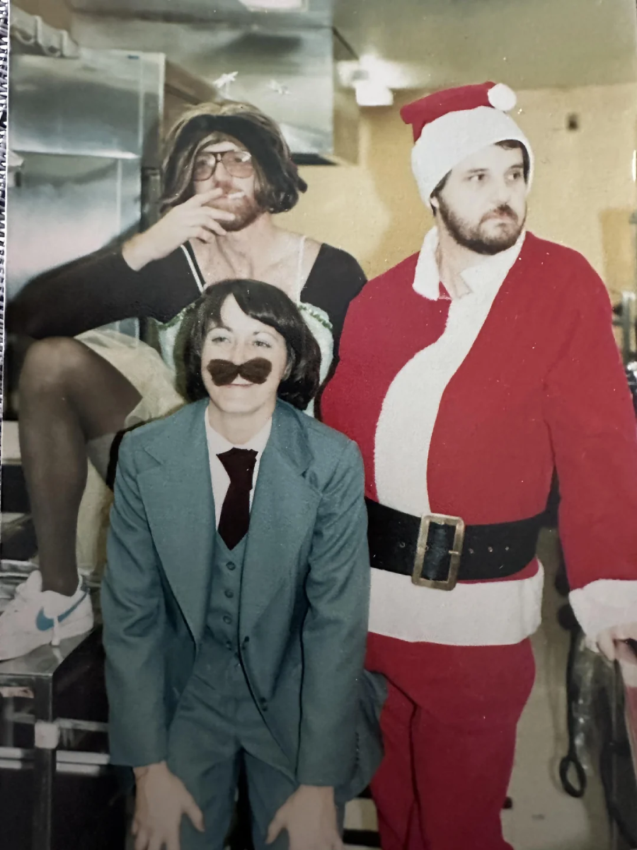 Three adults in costumes pose indoors: one in a Santa suit and hat, one in a blue suit with a fake mustache, and one in a wig, glasses, and casual clothes, smoking a cigarette. The background appears to be a kitchen.