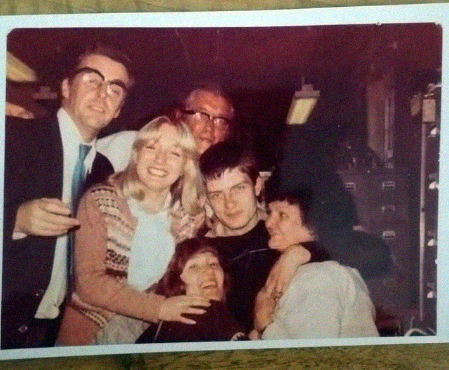A group of six people, smiling and hugging closely together indoors, posing for a photo. The background appears to show shelves or filing cabinets. The image has a warm, vintage tone.