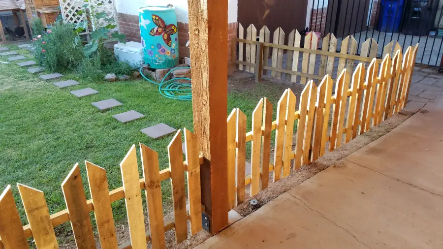 A wooden picket fence separates a grassy yard with stepping stones, flowers, and a butterfly-painted rain barrel from a concrete patio area. Sunlight casts shadows on the fence and ground.