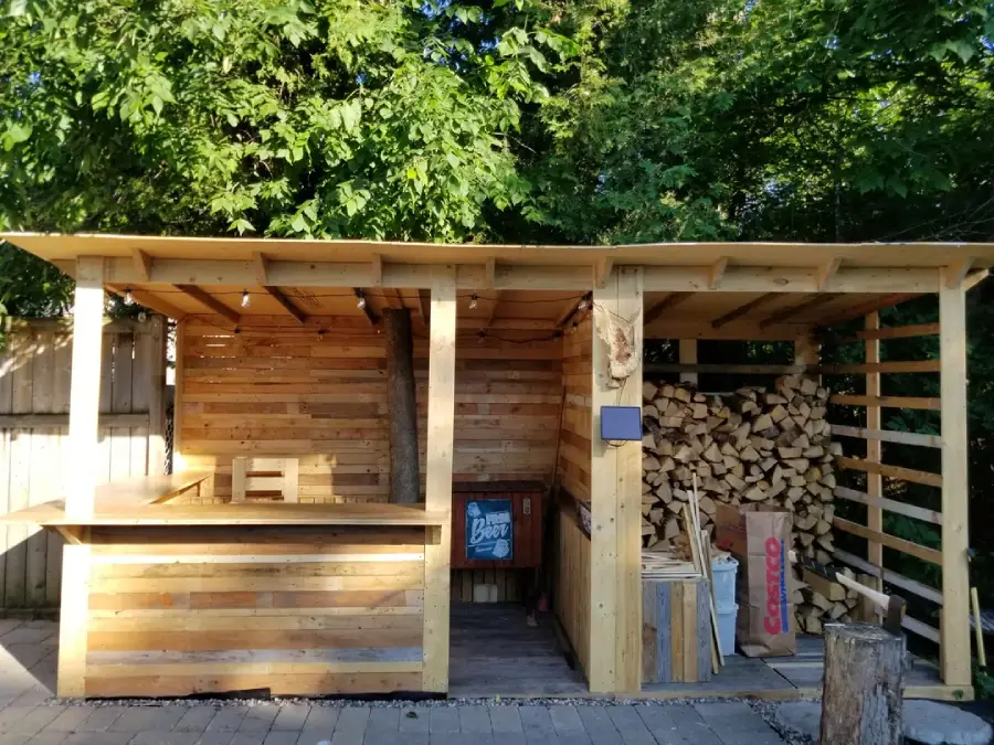 A rustic outdoor bar made of wooden pallets with a counter, barstool, partial roof, and a large stacked pile of firewood on the right, surrounded by green trees and sunlight.