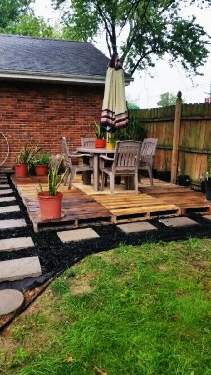 A small wooden patio with four chairs, a table, and a closed umbrella sits in a fenced backyard. Potted plants decorate the space, and a stone path leads to the patio, bordered by mulch and lawn.