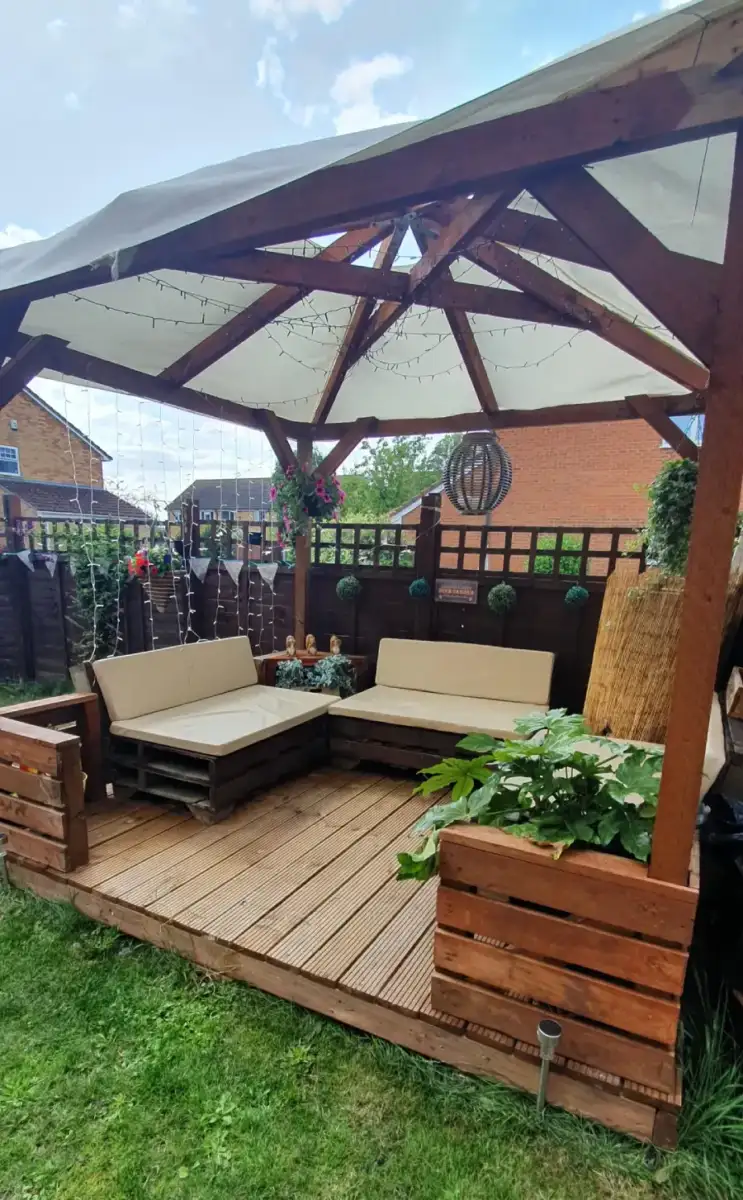 A wooden gazebo with a white canopy covers a cozy outdoor seating area with beige cushions, surrounded by plants and string lights. The space sits on a wooden deck in a grassy backyard.
