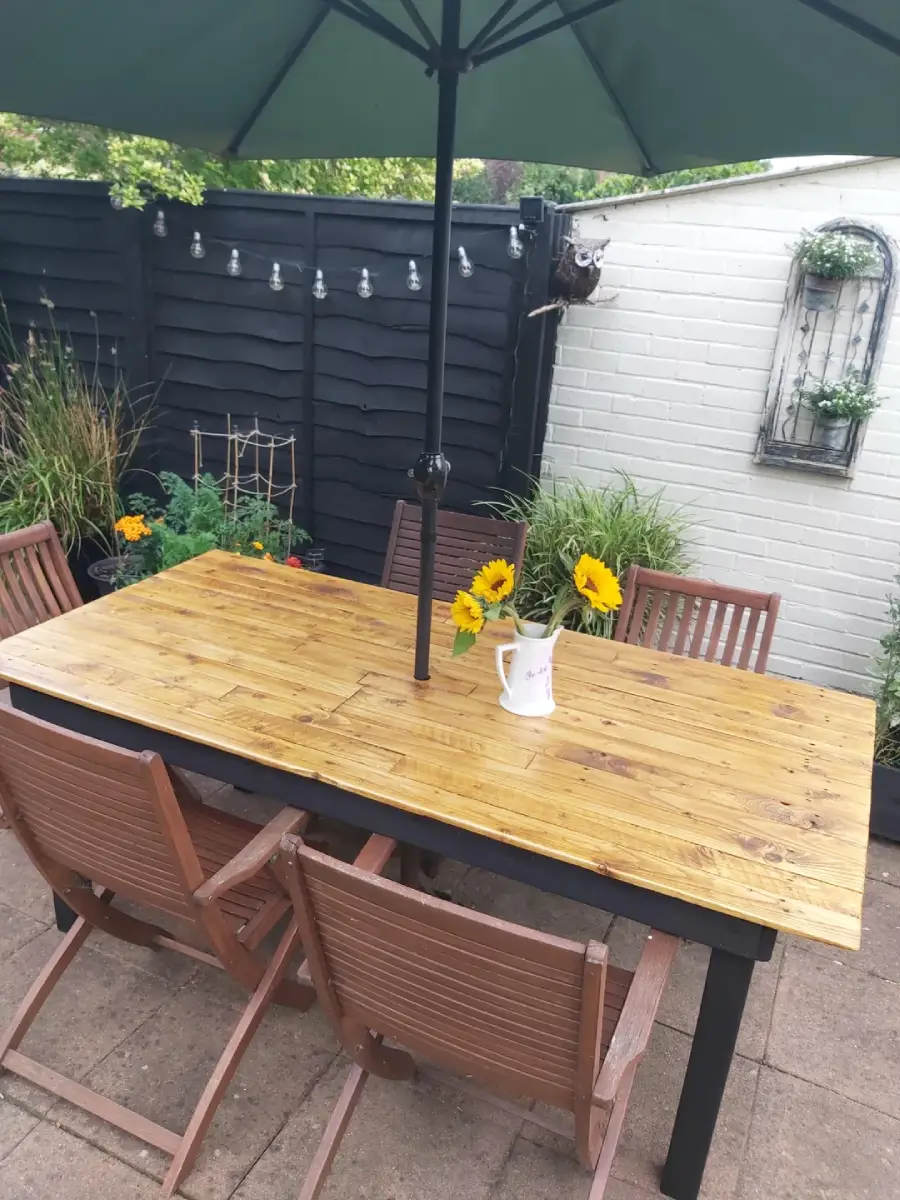 A wooden outdoor table with four chairs sits on a patio under a large green umbrella. A white jug with sunflowers is on the table. Surrounding plants and a white brick wall with hanging decor are in the background.