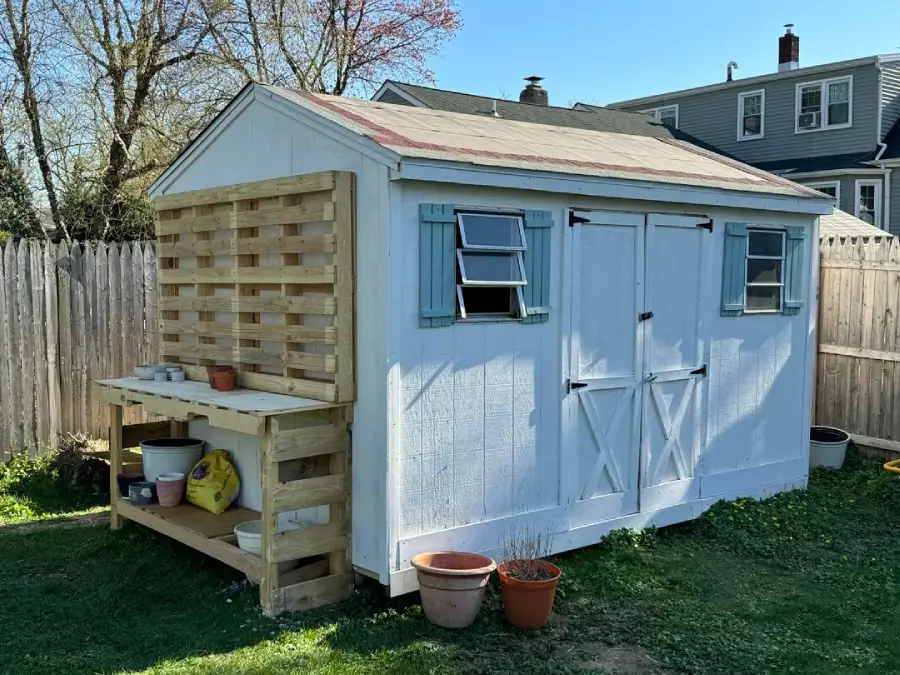A white shed with blue shutters stands in a grassy yard. A wooden potting bench with gardening supplies is attached to one side, and several potted plants sit on the ground nearby. A wooden fence and houses are in the background.