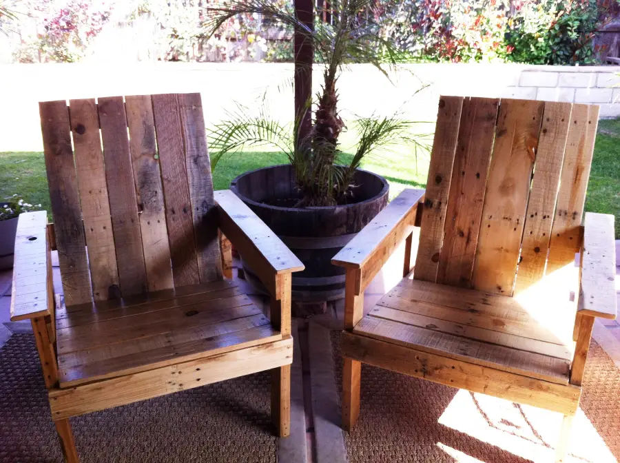 Two wooden chairs made from pallets sit side by side on an outdoor patio, with a potted plant behind them and a grassy yard in the background. Sunlight casts shadows on the chairs and floor.