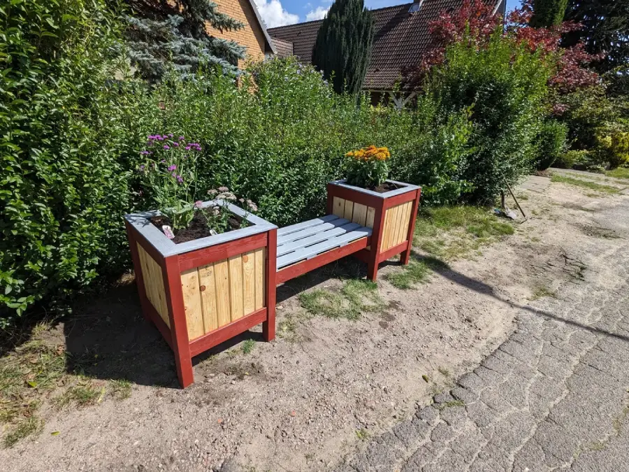 A wooden bench with planter boxes on each end, containing blooming flowers, sits on a sandy area beside a stone path with green shrubs and houses in the background.