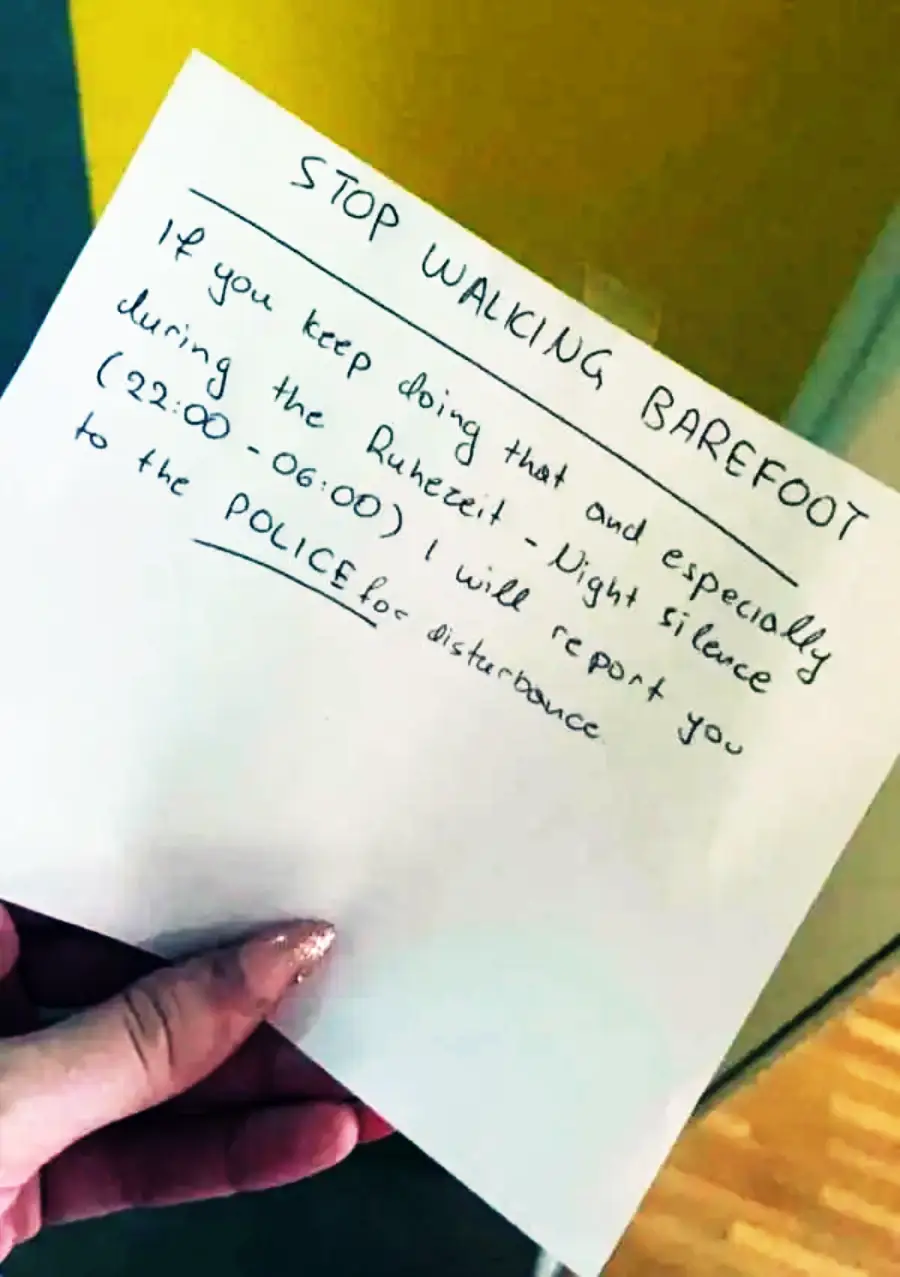 A hand holds a note that reads: "STOP WALKING BAREFOOT. If you keep doing that and especially during the Ruhezeit - Night silence (22:00-06:00), I will report you to the POLICE for disturbance.