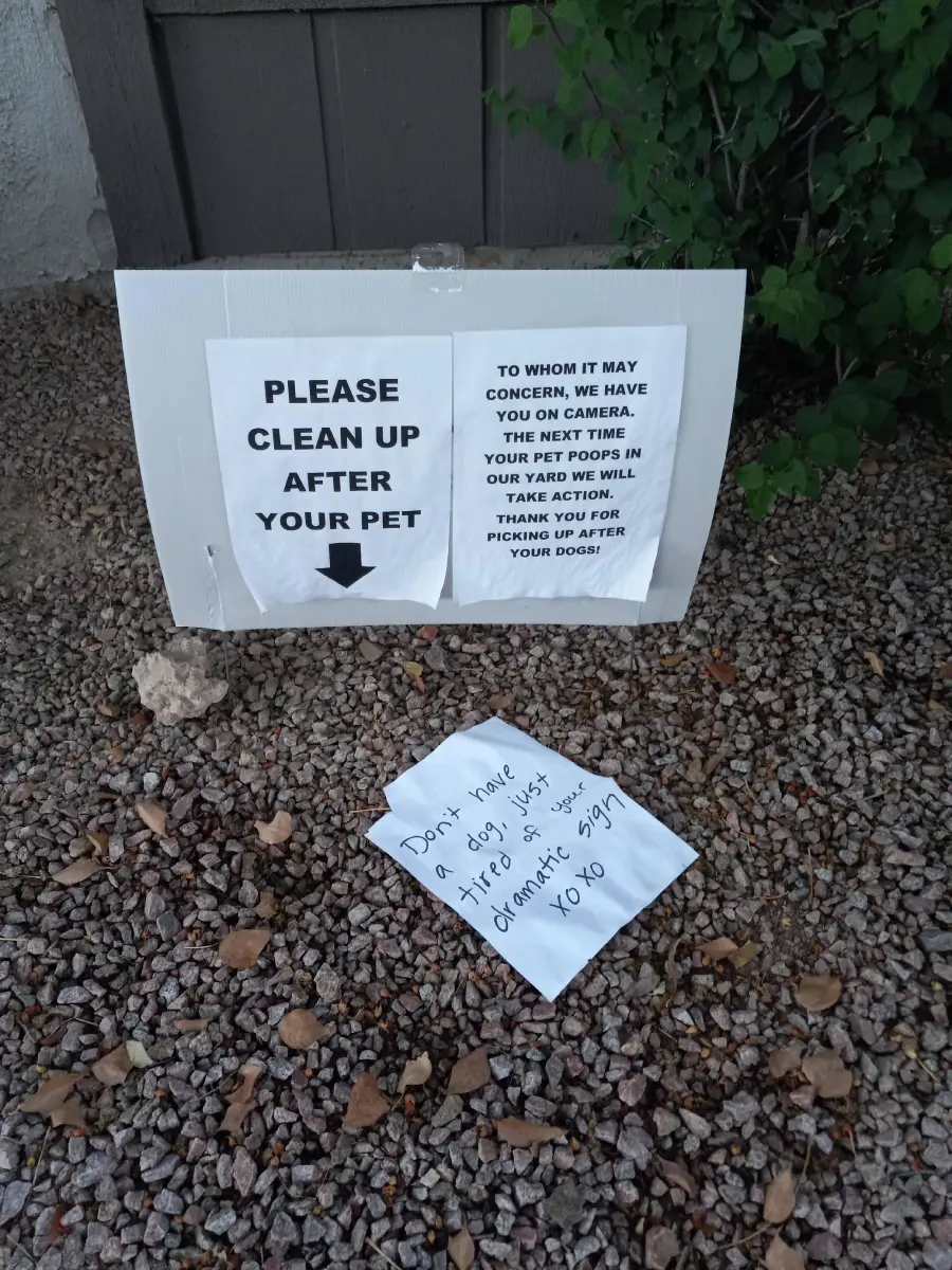 A yard sign reads “PLEASE CLEAN UP AFTER YOUR PET,” with a printed note warning about cameras. In front, a handwritten note says, “Don’t have a dog, just tired of this sign. Dramatic.” Both sit on gravel by a bush.
