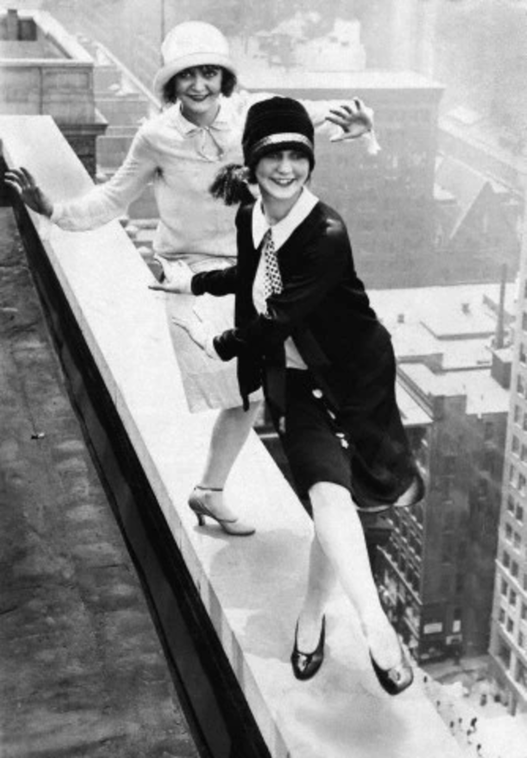 Two women dressed in 1920s fashion smile and pose on the edge of a tall building's rooftop, with a cityscape visible far below them.