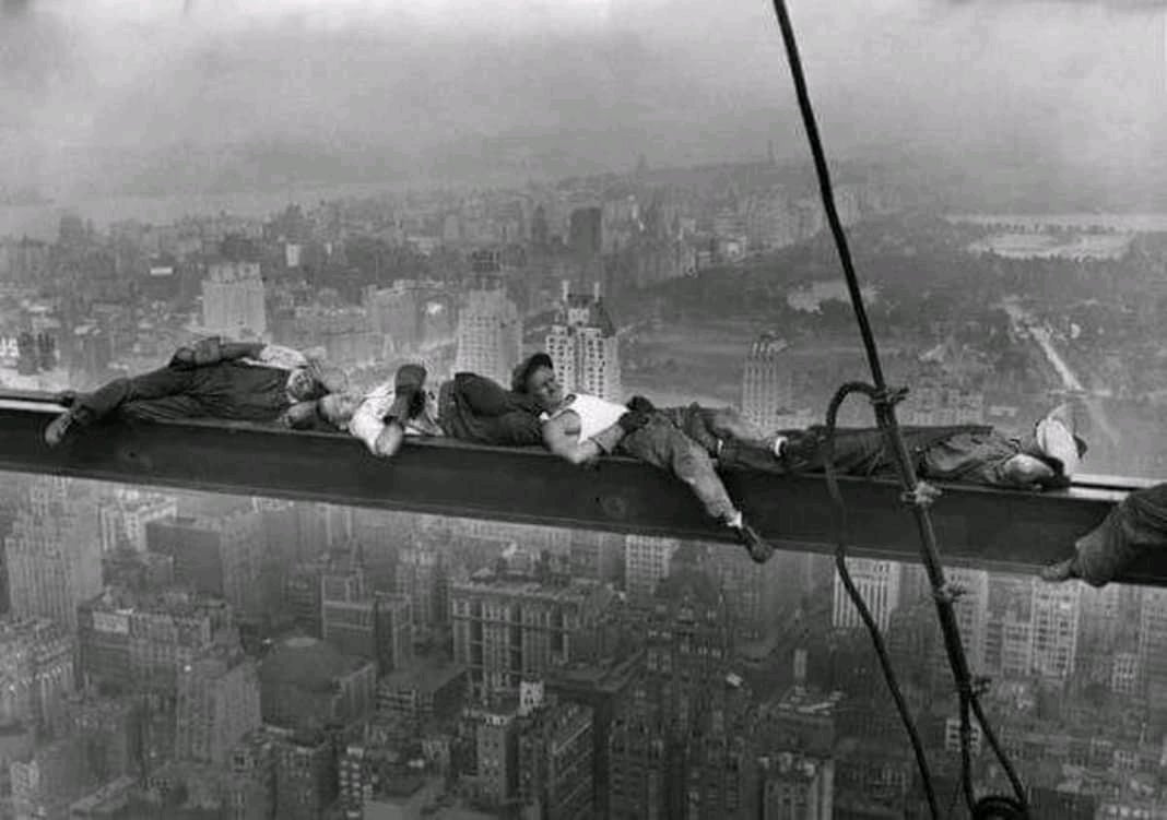 Four construction workers lie on a steel beam high above a city skyline, seemingly napping or resting, with buildings and a park visible far below in the background.