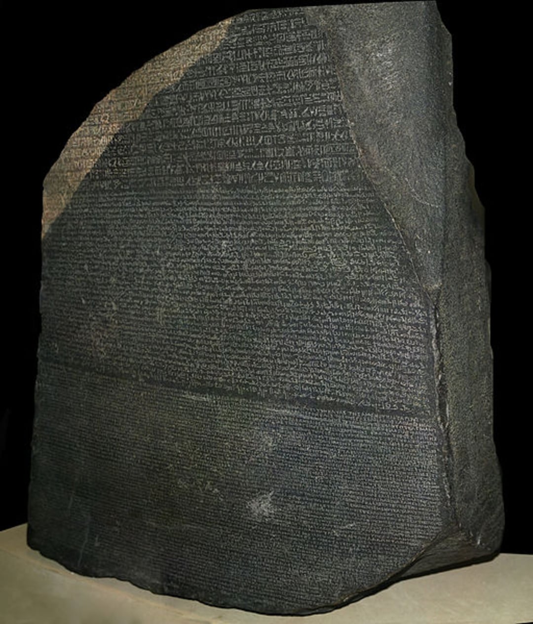 A large, dark stone slab featuring inscriptions in three different scripts, known as the Rosetta Stone, standing upright against a black background.