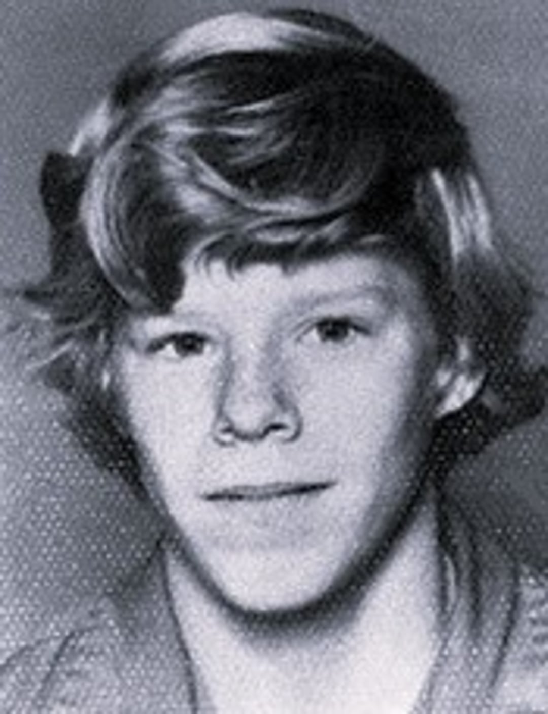Black and white portrait of a young person with light-colored, wavy hair, looking at the camera and smiling slightly. The background is plain and textured.