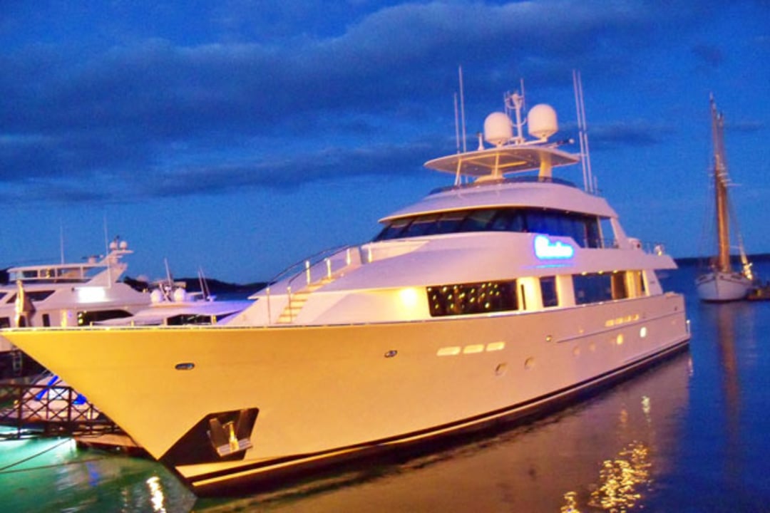 A large white luxury yacht docked at a marina during twilight, with soft lights glowing on the yacht and reflections shimmering on the calm water. Other boats and a sailboat are visible in the background under a blue sky.