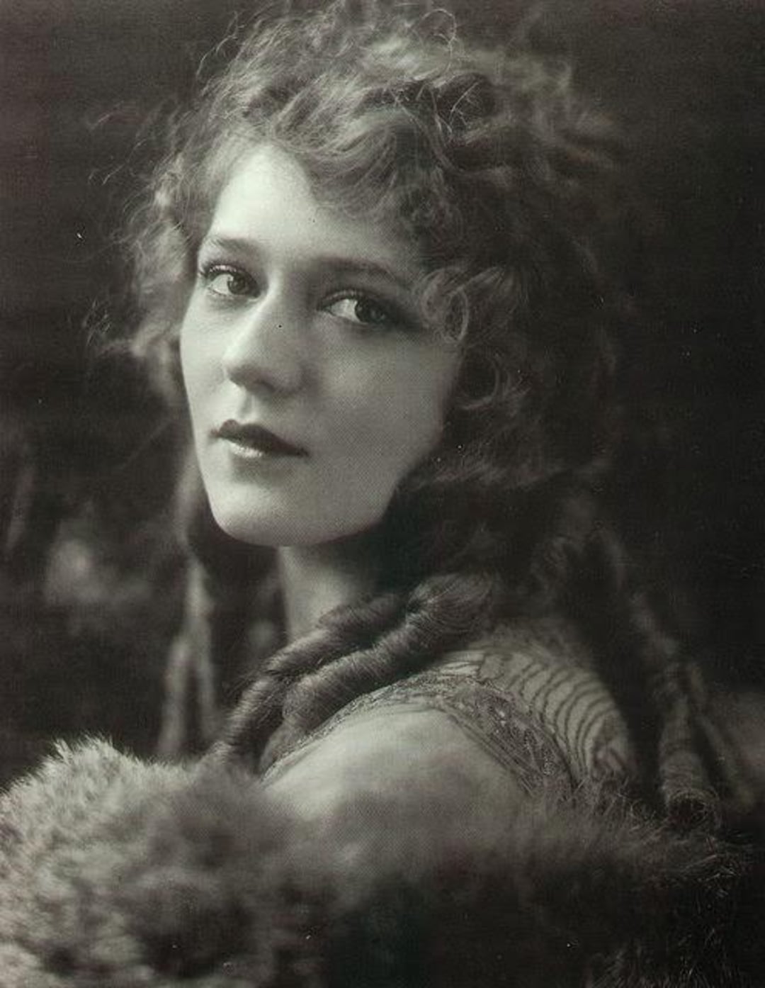 A black-and-white portrait of a young woman with wavy hair styled in loose curls. She looks slightly over her shoulder toward the camera, wearing a textured top and a fur-like wrap, with soft lighting highlighting her face.