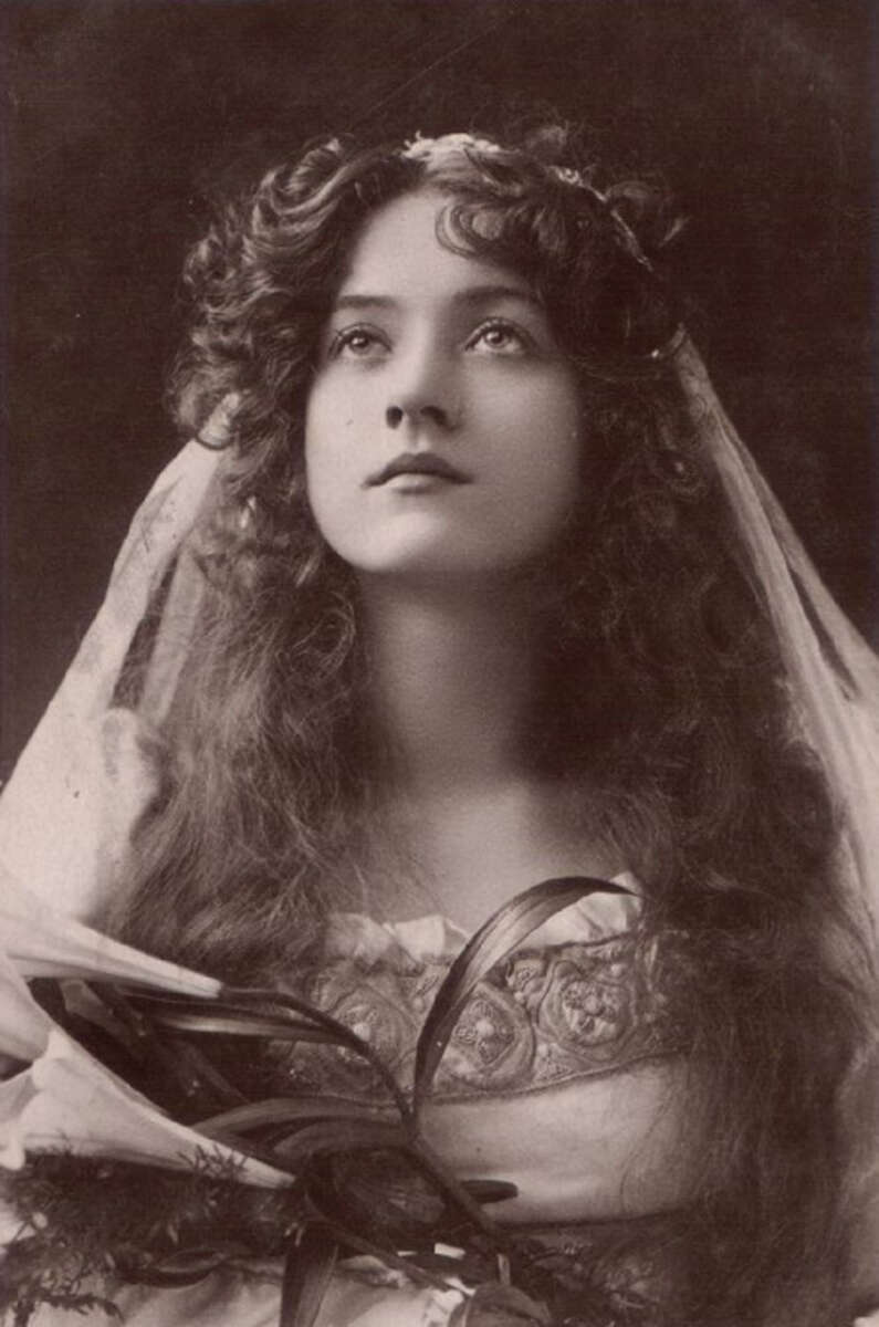 A vintage black-and-white portrait of a young woman with long, wavy hair, wearing a veil and decorative dress, gazing slightly upward with a thoughtful expression. She holds a bouquet of flowers with ribbon.