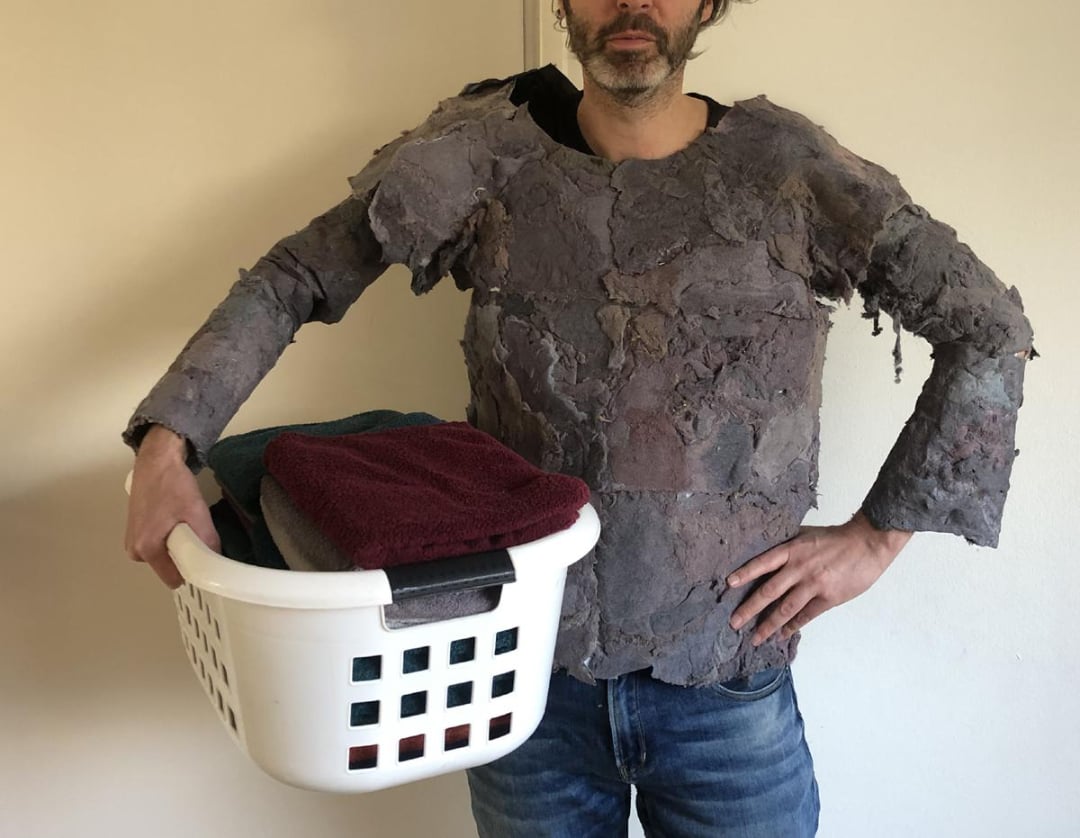 A man wearing a homemade, rough-textured costume that resembles stone or rock holds a laundry basket filled with folded towels against a plain wall.