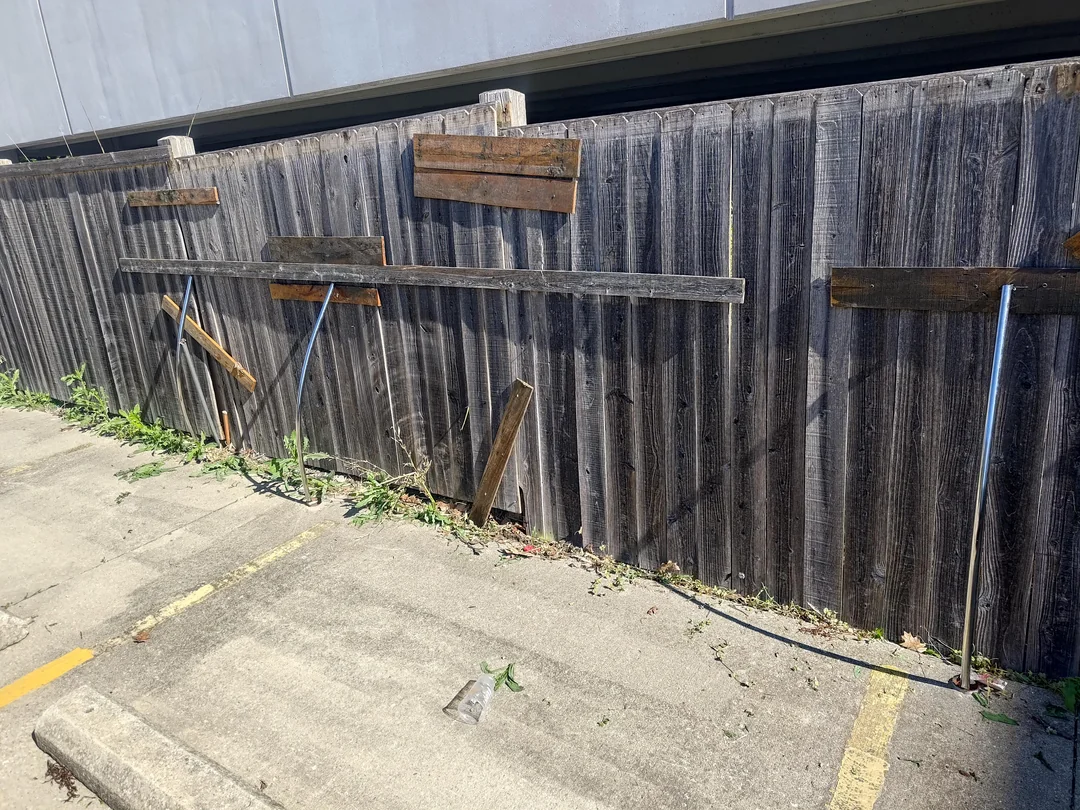 A damaged wooden fence with several metal poles leaning against it and wooden planks haphazardly nailed for support, next to a concrete parking area with yellow lines and some weeds growing along the fence.