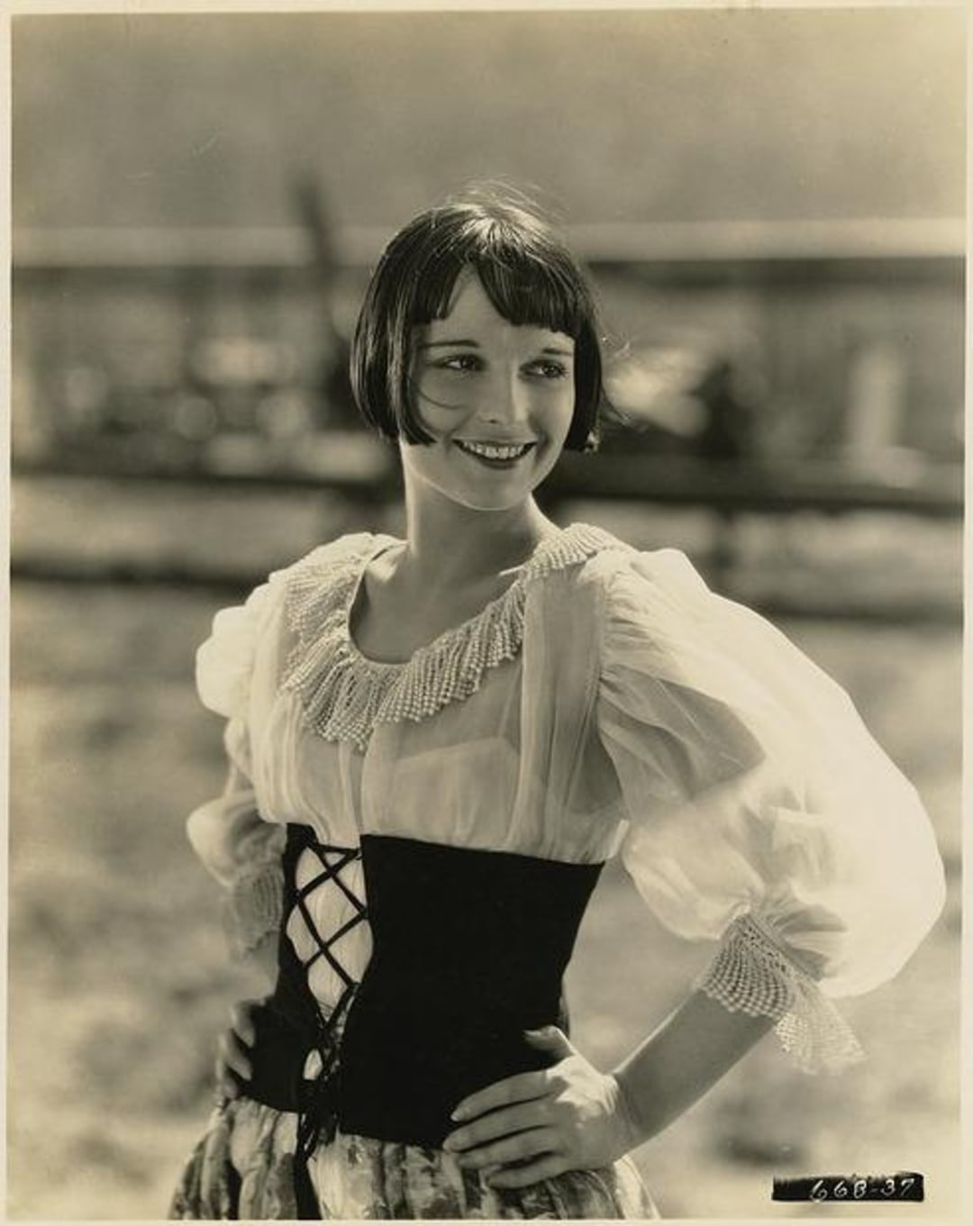 A woman with short, dark bobbed hair smiles while posing outdoors. She wears a sheer, puff-sleeved blouse with a lace-up corset and has her hands on her hips, exuding a cheerful and confident demeanor.