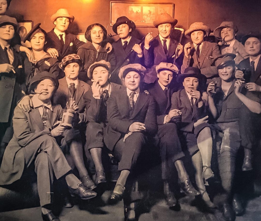 A group of women in suits and hats sit and stand closely together, many holding cigarettes and drinks, smiling and posing for the camera in a vintage, sepia-toned setting.