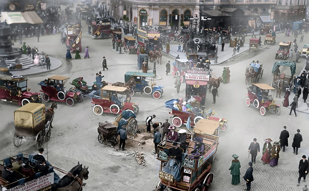 A busy early 1900s city street filled with horse-drawn carriages, vintage cars, double-decker buses, and pedestrians in period clothing; light fog or smoke lingers above cobblestone roads.