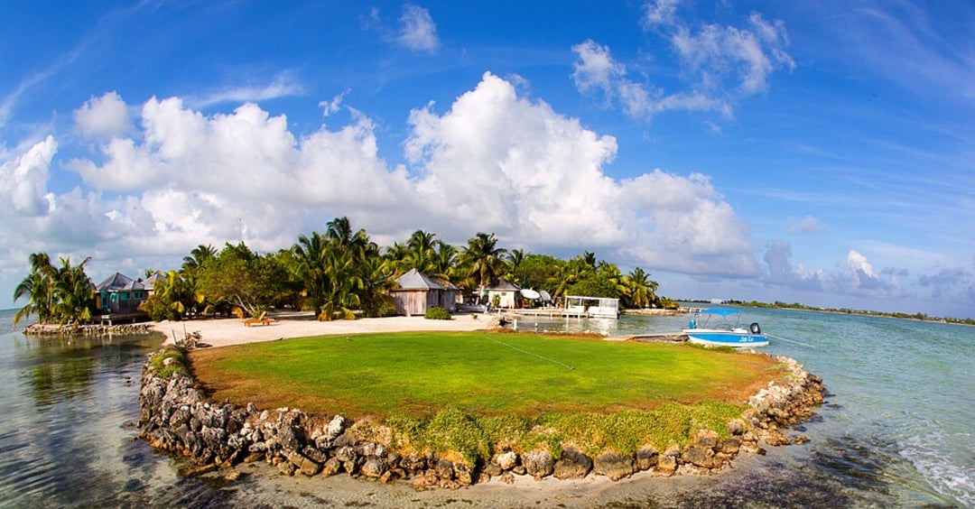 A small tropical island with lush palm trees, a few buildings, green grass, and a boat on clear turquoise water under a bright blue sky with scattered white clouds.
