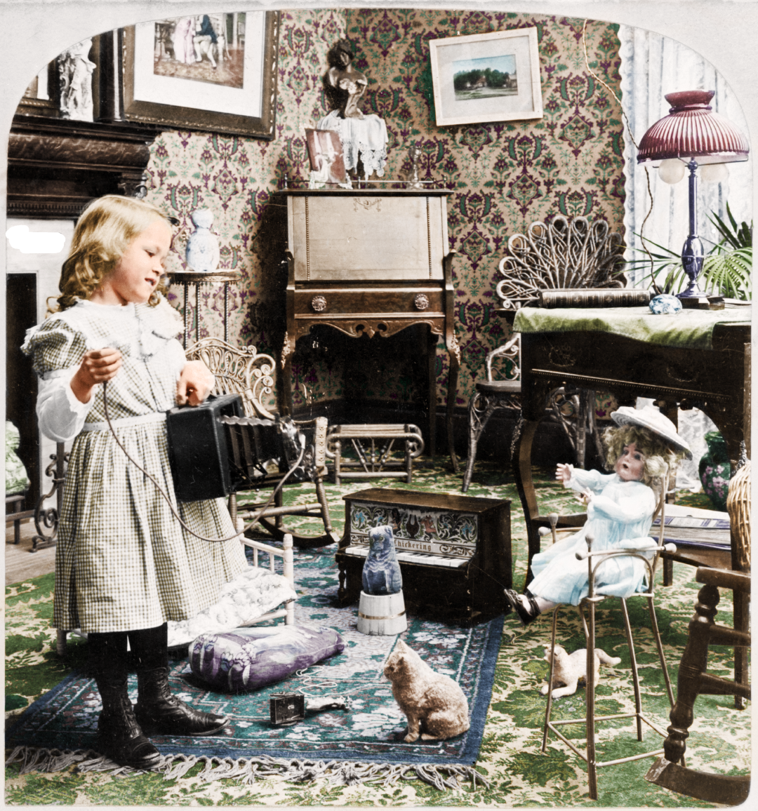 A young girl in a plaid dress pretends to photograph a doll seated on a chair in an ornate Victorian room filled with patterned wallpaper, antique furniture, and decorative items. A cat sits on the carpet watching.
