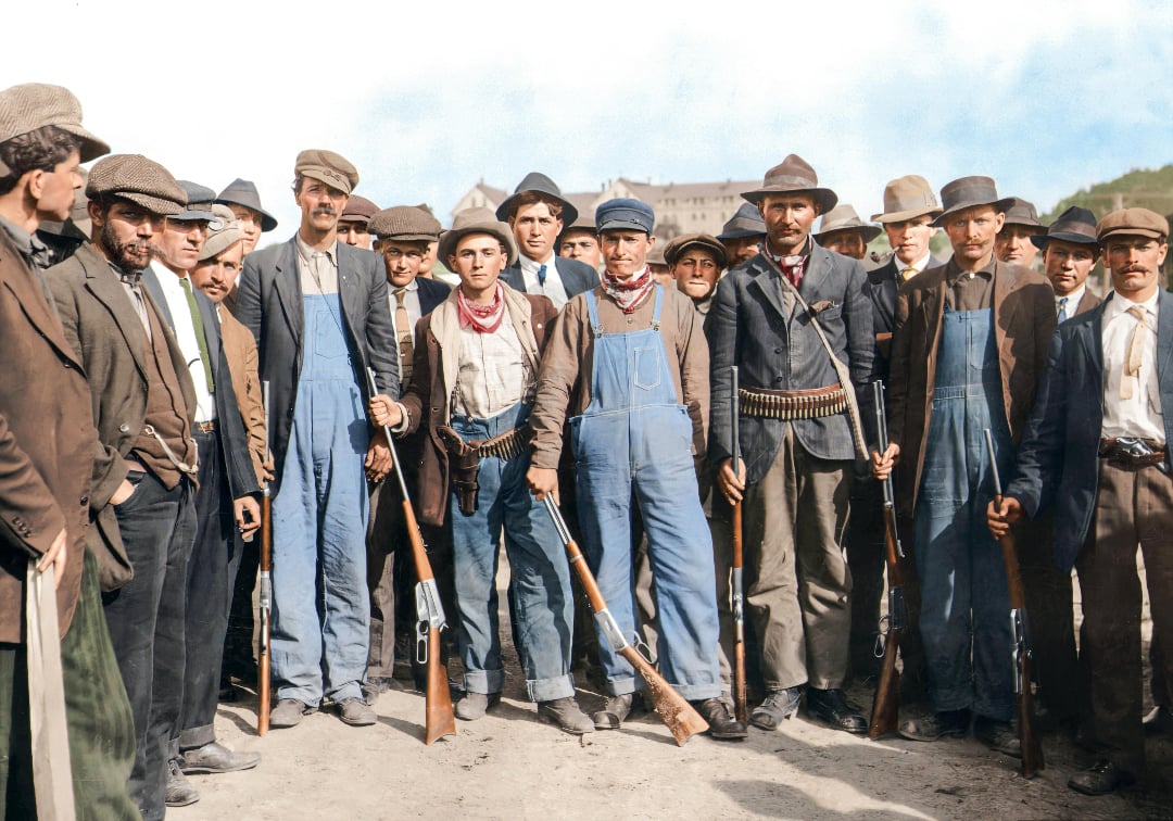 A group of men, many wearing hats, jackets, and overalls, stand together outdoors holding rifles. Several have ammunition belts across their chests. The mood appears serious, and a building is visible in the distant background.