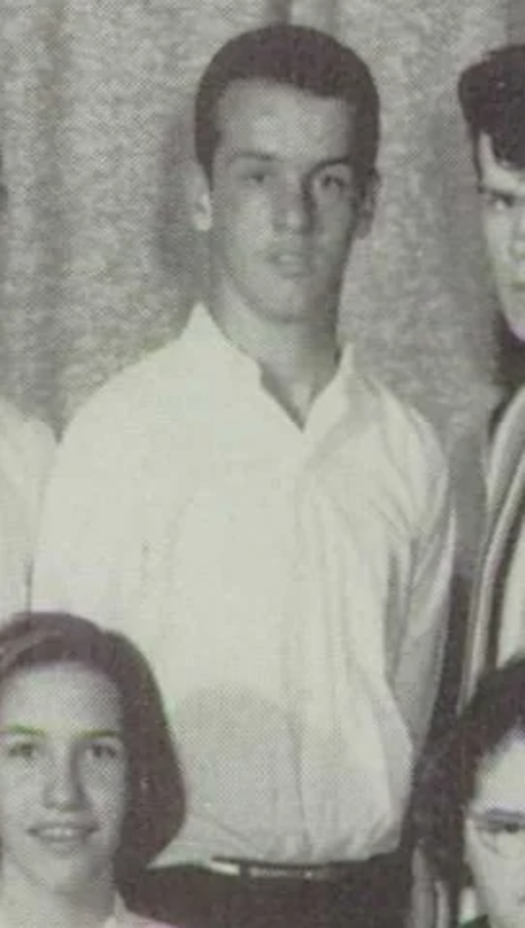 A black-and-white vintage photo shows a young man standing and looking at the camera, wearing a light-colored button-up shirt. Other people are partially visible around him.