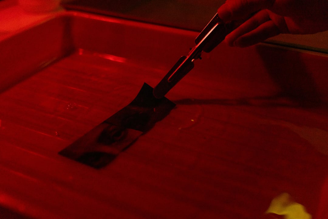 A hand uses tongs to hold a photographic print in a tray of liquid under red darkroom lighting, as an image slowly develops on the paper.