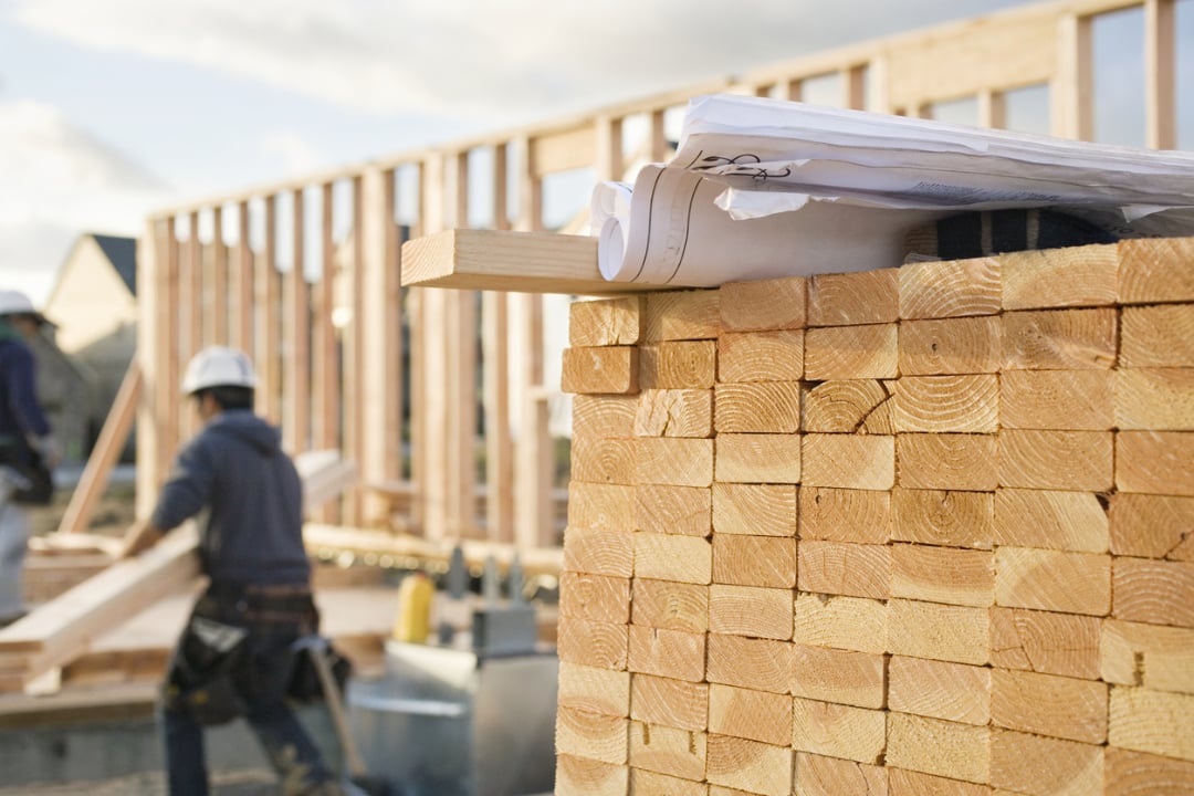 A stack of lumber and construction blueprints in the foreground, with construction workers and a partially built wooden frame of a house in the background.