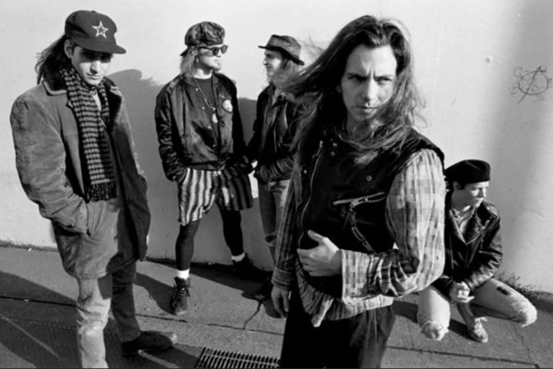 Five men in grunge-style clothing stand and crouch against a plain wall. One stands in front, looking toward the camera with long hair, while the others interact in the background, wearing jackets, hats, and sunglasses.