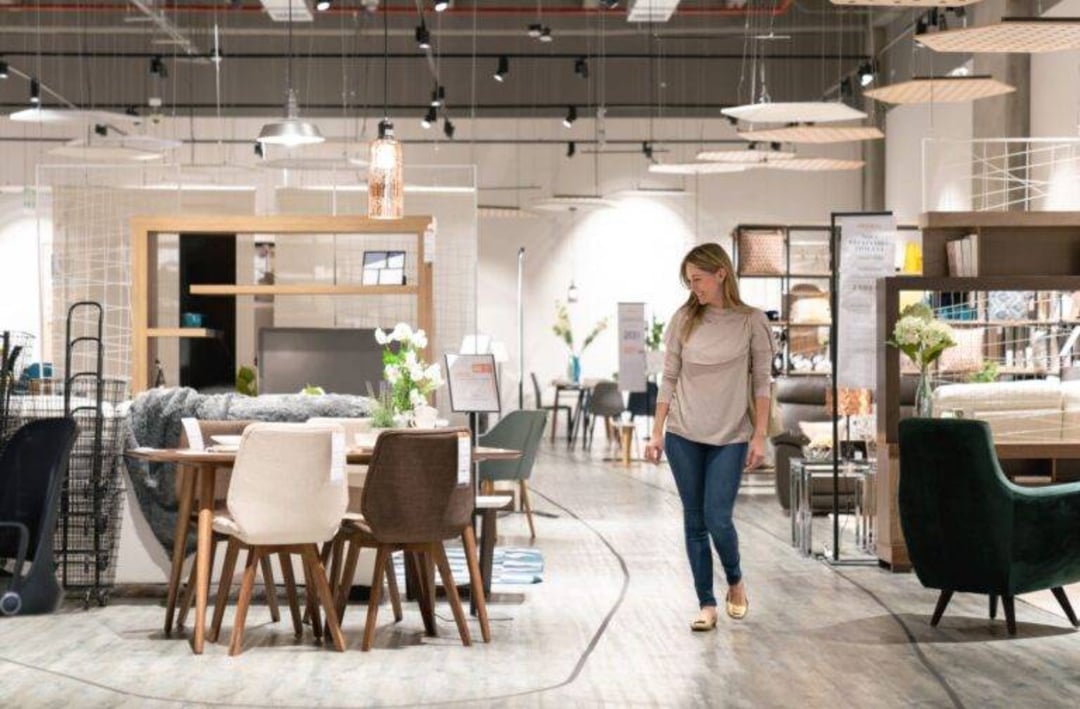 A woman walks through a modern furniture store, surrounded by dining tables, chairs, and home decor under bright lighting. The store has a spacious, organized layout with contemporary furnishings on display.