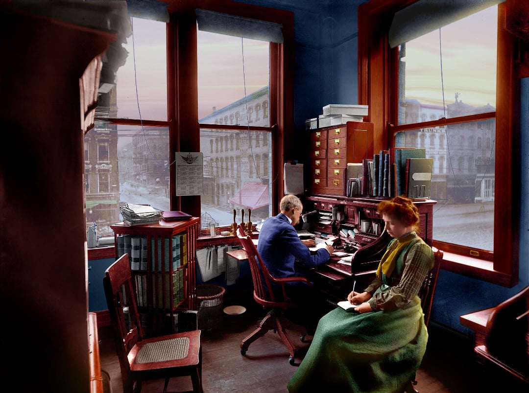 A man and woman sit in an early 20th-century office with large windows overlooking a city street. The man works at a desk covered in papers, while the woman sits nearby, writing in a notebook.