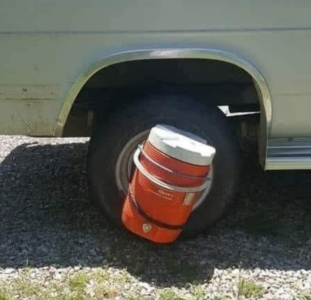 A large orange water cooler is strapped to a vehicle tire as if it were a wheel, replacing the usual tire, with the van parked on a gravel surface.