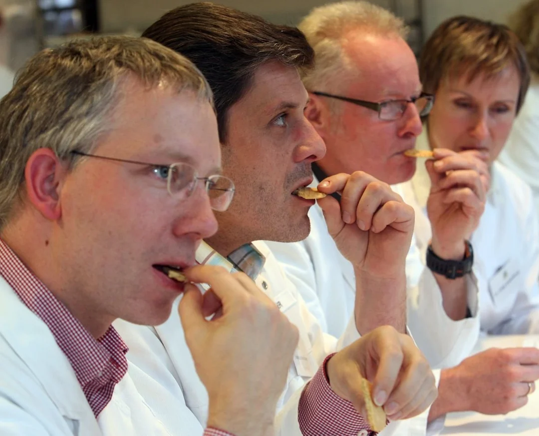 Four people wearing white lab coats sit in a row, closely tasting crackers. They appear focused and thoughtful, possibly evaluating the food as part of a taste test or scientific experiment.