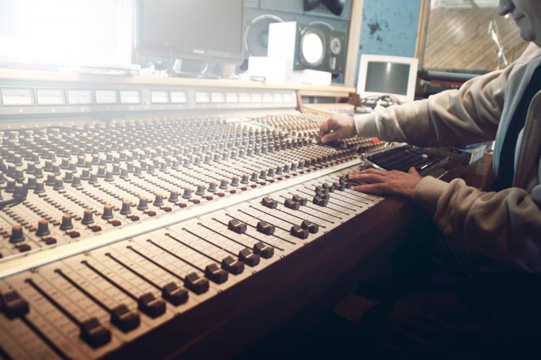 A person adjusts controls on a large audio mixing console in a recording studio, with various buttons, faders, and equipment visible on the desk.