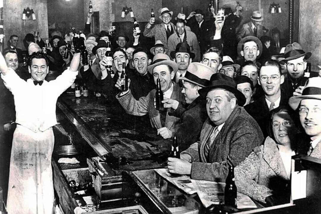 A large group of people in 1920s-style clothing cheer and raise bottles in a crowded bar, with a smiling bartender standing at the counter with arms raised in celebration.