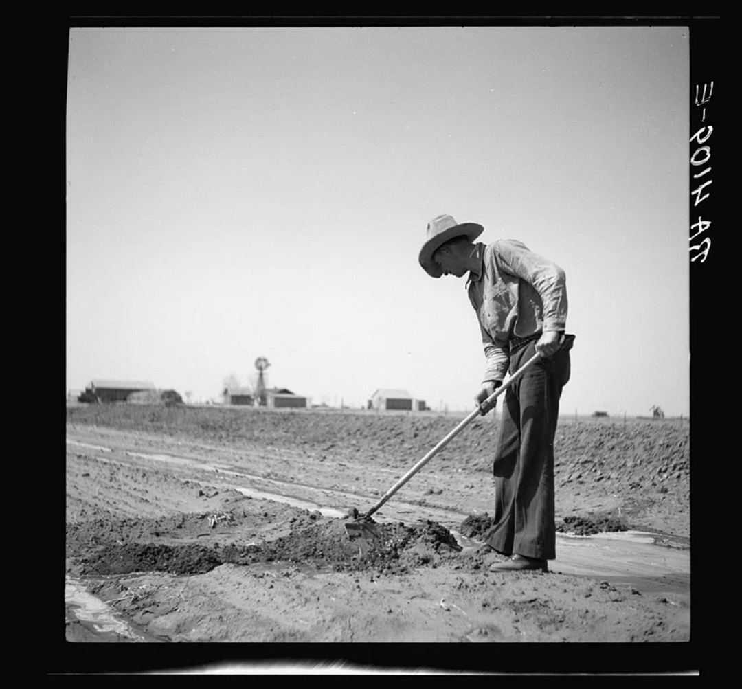 A man wearing a hat and work clothes uses a hoe to tend to a field on a farm. Farm buildings and a windmill are visible in the background under a clear sky.