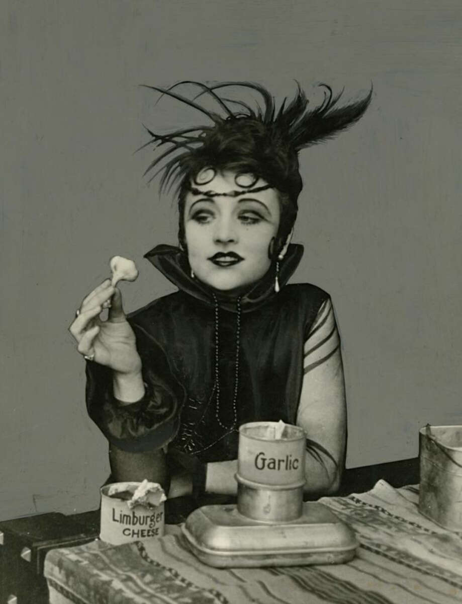 A woman in vintage attire with dramatic makeup and a feathered headpiece holds a clove of garlic. In front of her are tins labeled "Limburger Cheese" and "Garlic" on a table with a striped cloth.