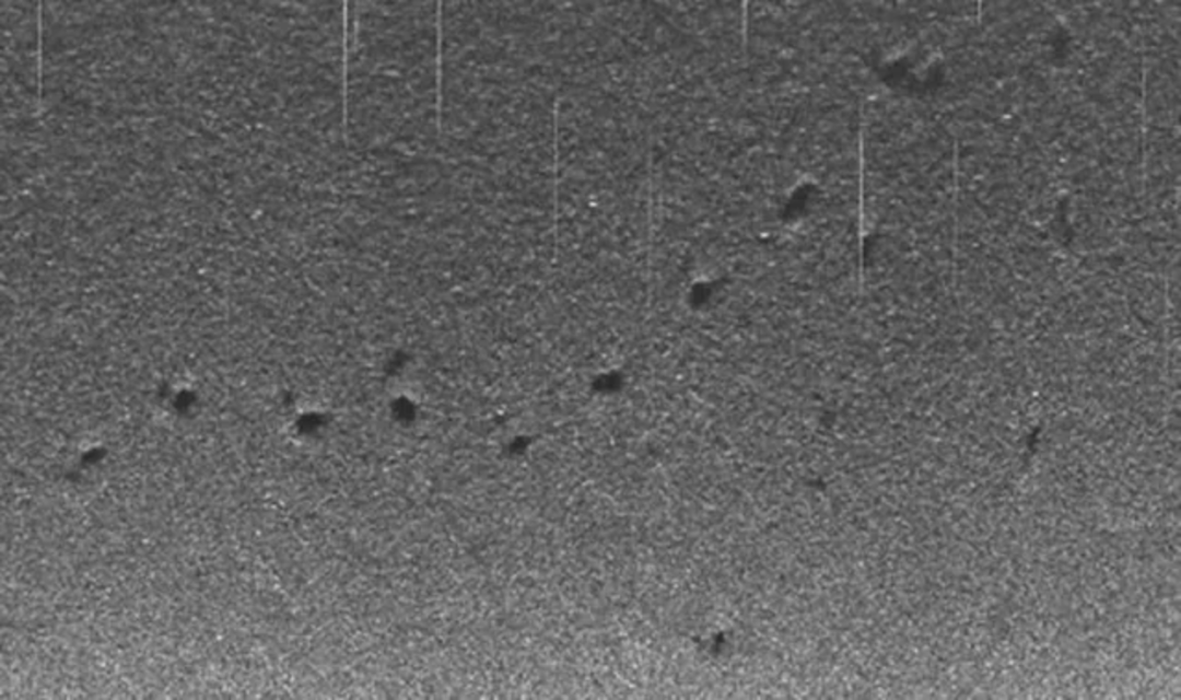 A grayscale image showing a grainy, textured surface with numerous small, dark, irregularly shaped craters scattered across it. Thin vertical lines stretch from the top, resembling scratches or data artifacts.