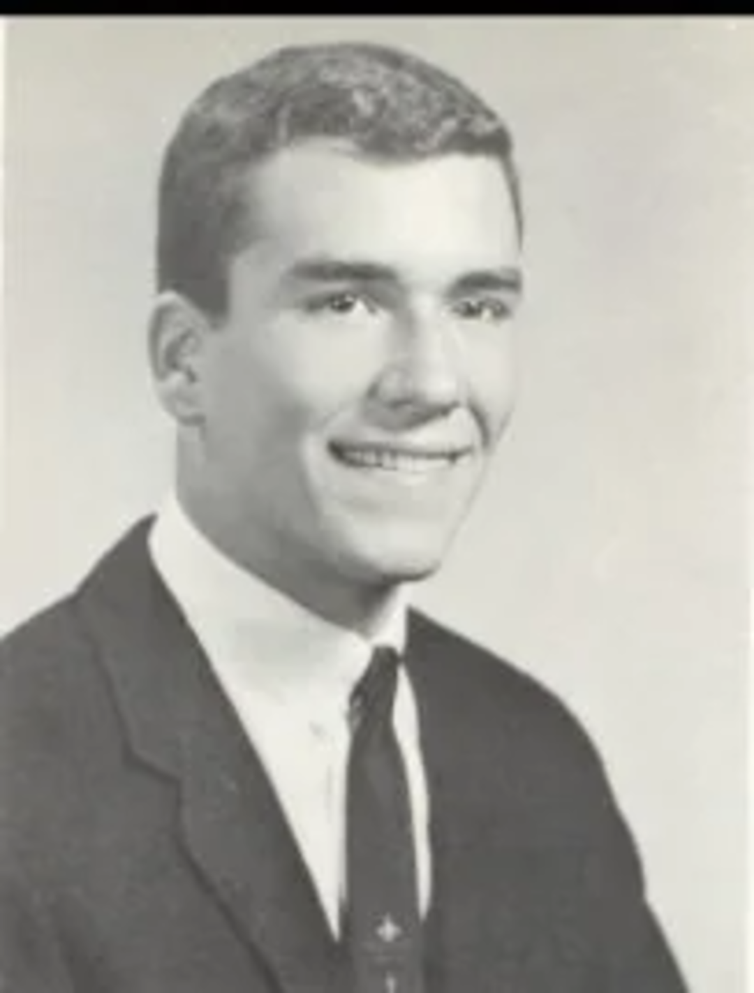 Black and white portrait of a young man in a suit and tie, smiling and looking slightly to the side, with short hair and a clean-shaven face, against a plain background.