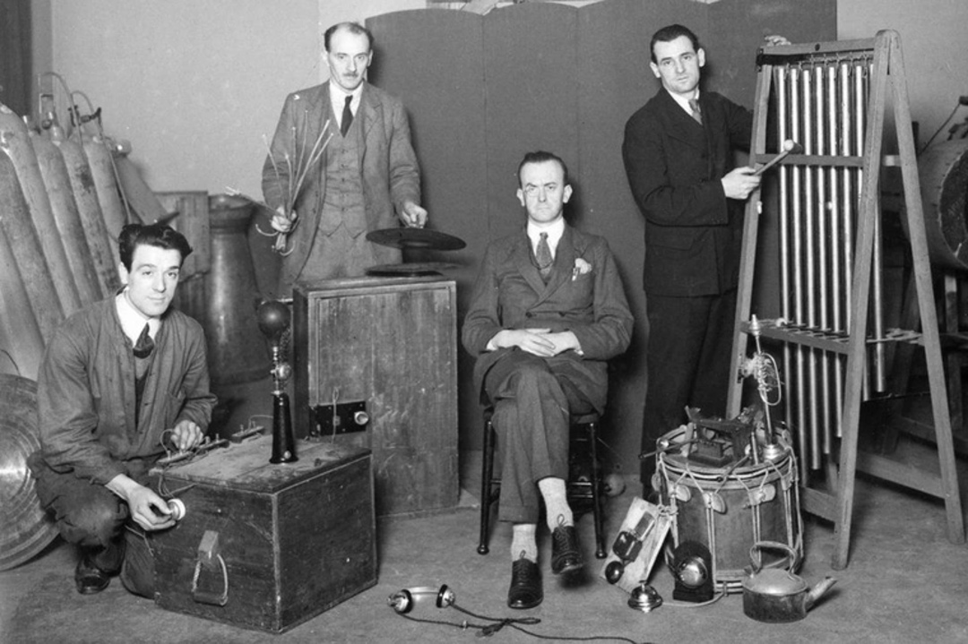 Four men pose with various old-fashioned sound effects equipment, including chimes, drums, and other devices in a vintage black and white setting, suggesting an early sound production or foley studio.