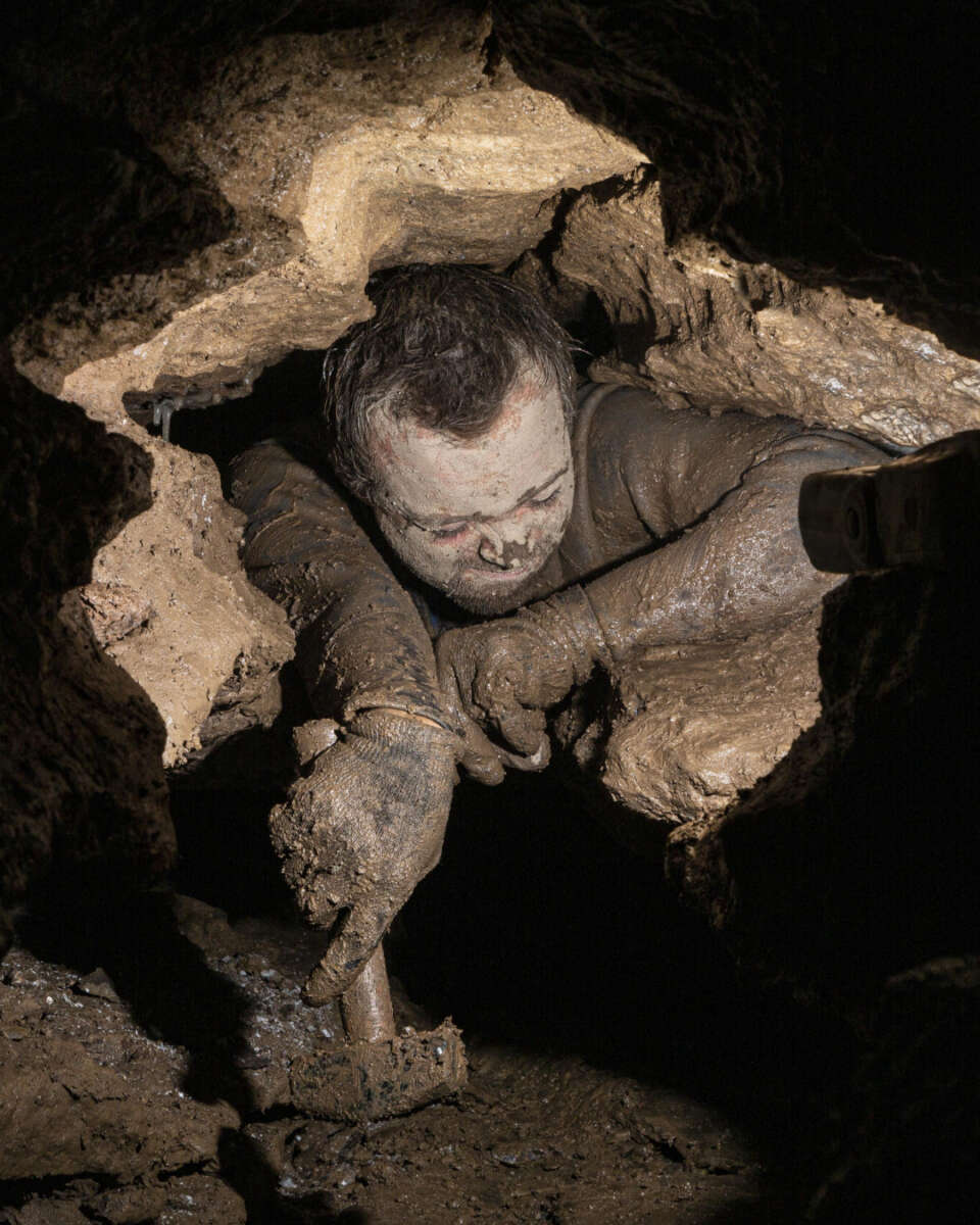 A person covered in mud squeezes through a narrow, rocky cave passage, using their arms to navigate the tight space. Dim light highlights their determined expression and the rough cave walls.