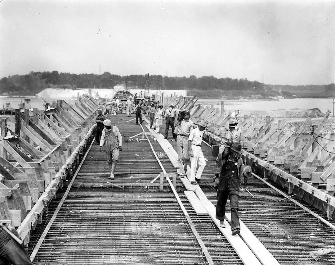 Black-and-white photo of construction workers building a bridge; they walk on wooden planks and metal framework, with trees and water visible in the background.