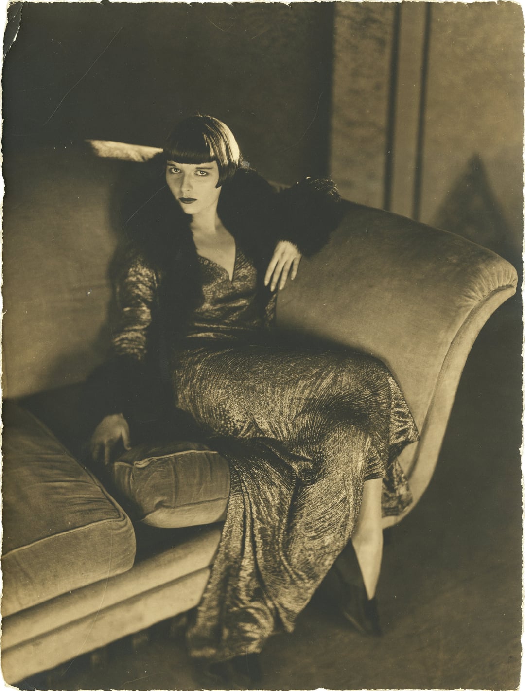 A woman in a shimmering gown and fur-trimmed sleeves lounges on a large, plush sofa. She has a short bob haircut with bangs and gazes intently at the camera, exuding a sense of vintage glamour and elegance.