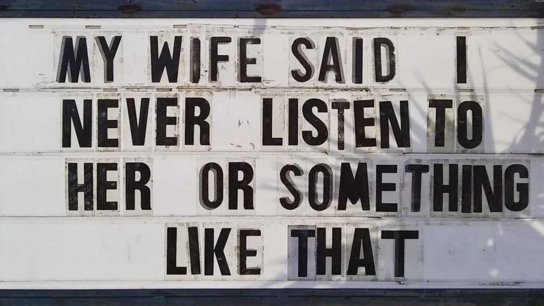 A sign reads, "MY WIFE SAID I NEVER LISTEN TO HER OR SOMETHING LIKE THAT" in bold black letters on a white background. There are faint shadows of tree branches on the sign.