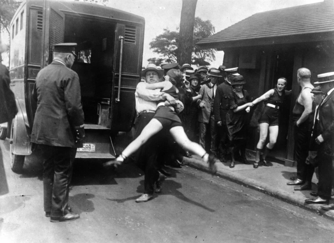 A police officer carries a woman in a swimsuit toward a police van as a crowd, including officers and swimmers in suits, watch during a public incident, likely from the early 20th century.