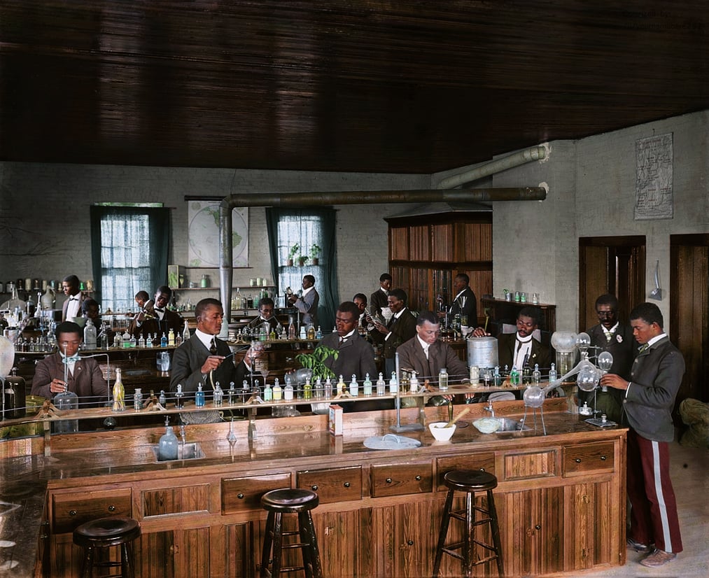 A group of Black men in suits work with glassware and chemicals in a large, early 20th-century science laboratory with wooden benches and shelves lined with bottles. Natural light enters through tall windows.