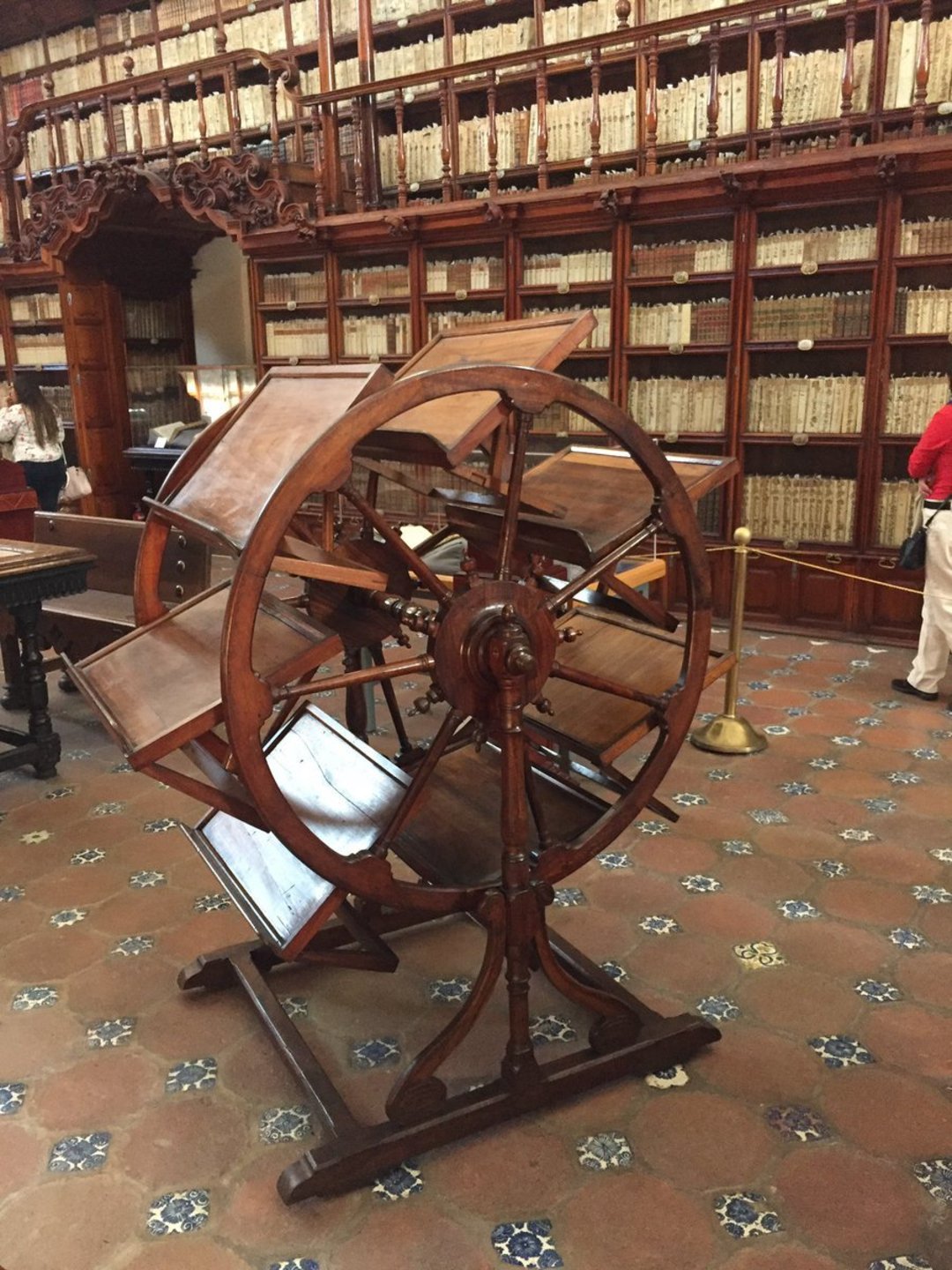 A large wooden bookwheel with multiple angled shelves stands in the center of an ornate library lined with tall bookshelves filled with old books. The floor is tiled, and a few people are visible in the background.